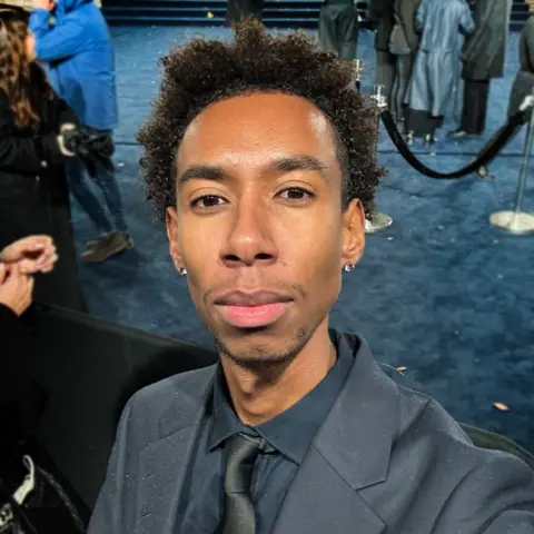 Coldest Creative A smartly dressed young man poses for a selfie in front of a blue carpet. Velvet ropes for VIP areas and people milling about as they await the arrival of important guests are visible.