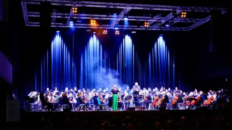 City of Wolverhampton Council A large orchestra is seated on a dramatically lit stage with purple and blue lighting and stage smoke. Musicians are in traditional orchestral formation with strings at the front and wind and percussion instruments behind. The stage is framed by dark curtains.