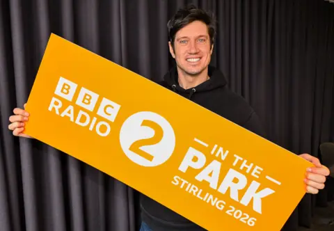 BBC Vernon Kay, a man in his 40s with dark hair, holds up a sign which says BBC Radio 2 in the Park Stirling 2026