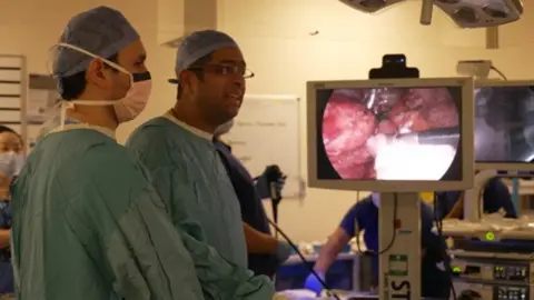 Two men in blue scrubs and masks looking at a screen displaying organs