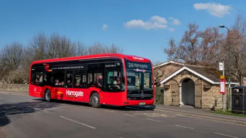 A red bus which says Harrogate on the front, pulling into a stone bus stop which is an attractive small building with an arched doorway and open gates. 