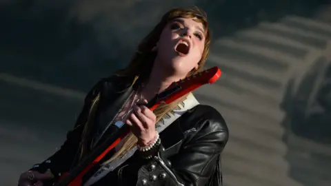Getty Images A woman singing on stage. She has long brown hair and is wearing a black leather jacket and holding a red guitar