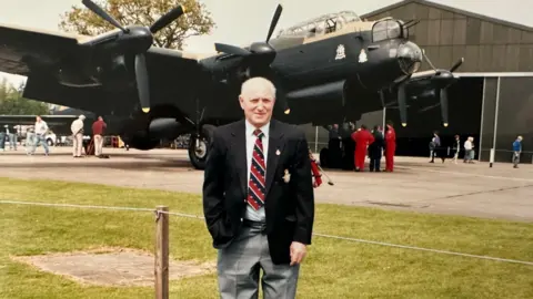 Ivor Taylor standing in front of a Lancaster bomber, which is on display at an airfield. He is wearing grey trousers, a white shirt, black blazer and a red and black striped tie.