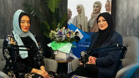 Two women sitting on grey chairs with a table that has a bouquet of flowers on top. The woman on the left is wearing a light blue hijab and a floral cardigan while the woman on the right is wearing a black hijab and navy blue blazer.