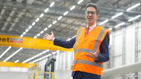 PA Media Energy secretary Ed Miliband stands smiling inside a large, modern-looking factory. He is wearing an orange and grey hi-vis jacket.