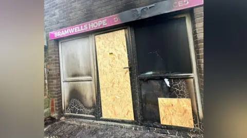 Bramwells Hope The front of a shop which has been badly damaged by fire. It has a pink sign which says Bramwells Hope and the door and one lower window have been boarded up. 