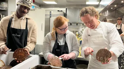 Chef Tom Kitchin, a curly-haired man in chef's whites, demonstrates opening the shell to a young man and woman in his kitchen. They wear dark aprons over their clothes and sous chefs work away in the background