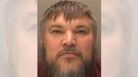 A police custody photo of a man's face taken from close range. He has short, greying brown hair and a greying beard.