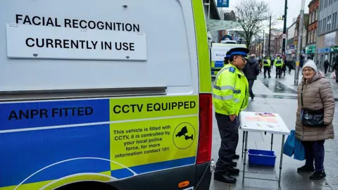 A Thames Valley Police community support officer pictured standing at a table with a woman dressed in a thick coat and woolly hat looking at the camera, with a Thames Valley Police van on the left.
