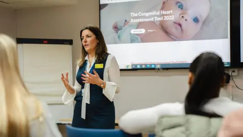 A woman with shoulder-length brown hair, wearing a white blouse with a sleeveless blue dress. She appears to be delivering a talk to a room of people. A screen behind her has a picture of a baby labelled "The Congenital Heart Assessment Tool 2022".