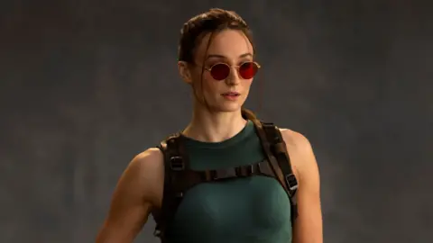 A photo shows Sophie Turner as Lara Croft. She wears red-tinted sunglasses, her hair tied back. She wears a green vest top and tight, thigh-length shorts. One of her fingerless-gloved hands rests on a pistol in a thigh holster.