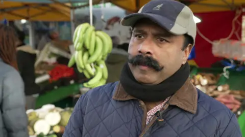 Jose Joseph, wearing a baseball cap, stands in front of his market stall, a large bunch of bananas hanging in the background
