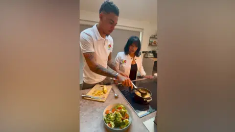 Zena Weeks Rogers on the left has his hands near a pan at the bottom of the image close to a chopping board and a dish. Weeks is on the right looking on.