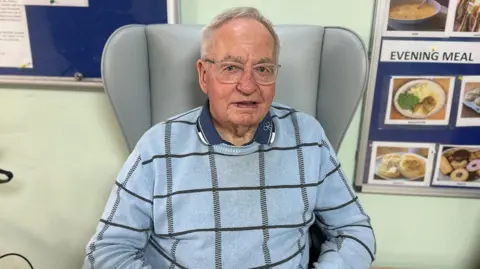 Alfred Fairclough is sitting in front on a chair in the lounge area. There are a number of notice boards behind him with newsletters and information for residents and their families. 