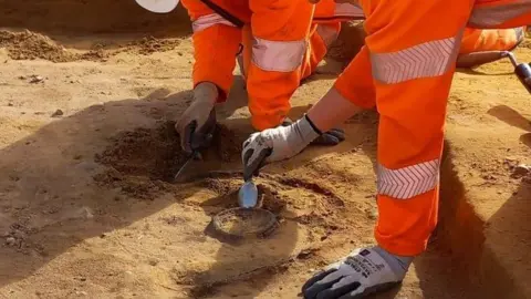 OCA A close-up of the arms of two archaeologists in hi-vis orange clothing, leaning into a trench and excavating two round dishes, one within the other. In right hands are trowels. The nearer archaeologist is using their trowel to reveal a silver dish and the far archaeologist is using their trowel to reveal a copper dish. 