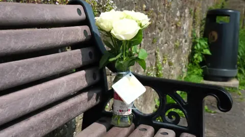 A small bunch of white roses left on a brown and black bench close to the River Ribble in Ribchester.