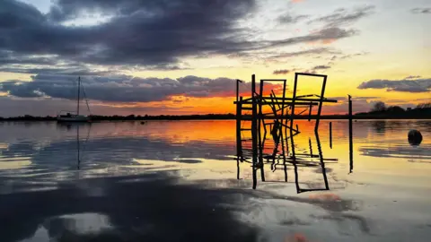 Simonbuk A vivid orange sunset over the harbour is reflected in the water. The sky has dark clouds above the orange/yellow glow. In the water you can see a sailing boat and a collapsed structure. 