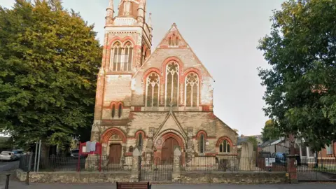 Google The exterior of a church building. There are trees on either side of it and the sky is blue.