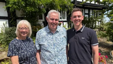 Three people stand in front of a tudor style black and white building. On the left is a woman with light blond hair, she is wearing glasses and a navy top. In the middle is a grey haired man with a pale blue and white patterned shirt and on the right is a younger man with short dark blond hair and a dark blue polo shirt.