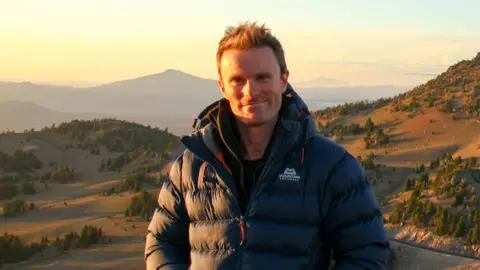 Supplied James stands on a large hill with trees and mountains in the background 