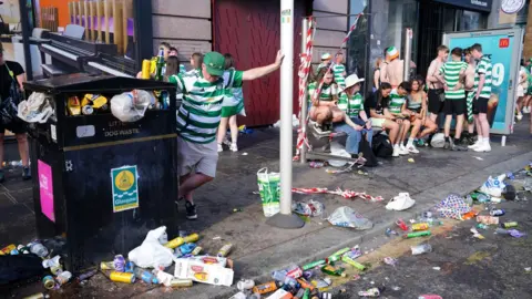 PA Media Celtic fans in Celtic shirts sitting at a bus shelter with no glass in it. They are surrounded by rubbish on the street and a fan leans on an overflowing black council bin