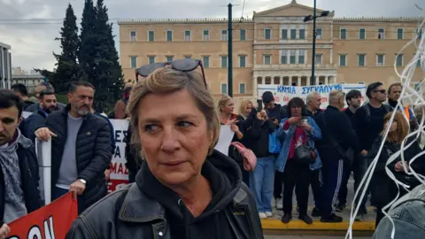 BBC/Kostas Koukoumakas A woman stands among protesters clutching white balloons which are out of the picture