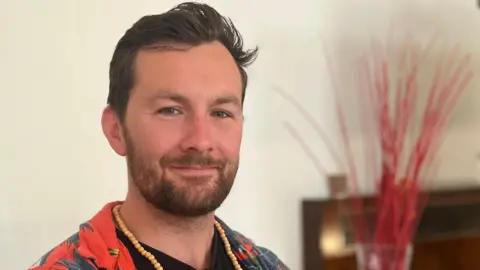 Aaron Mackenzie in a black T-shirt and a red Hawaiian shirt smiles at the camera. He has slicked-back black hair and is wearing a braided necklace.