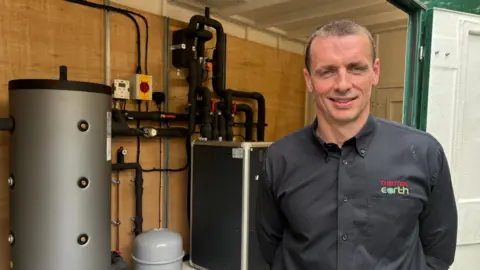 Nick Salini with short hair and wearing a black shirt, standing right of frame. Behind him is a heating system with black pipework, a compressor and boiler system.