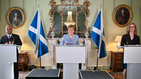 A bald man in a dark suit stood behind a rostrum and to his left is a woman in pale blue suit stood behind a rostrum, and to her left is a woman in a dark dress stood behind a rostrum