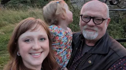 PA Media Cicely, pictured on the left, is smiling at the camera with a small child in the centre and her father on the right. She has red hair, cut into a fringe, and the child has blonde hair in pigtails and is wearing a multicoloured floral dress. Mr Berry is wearing a brown jacket or gilet with a t-shirt on. 