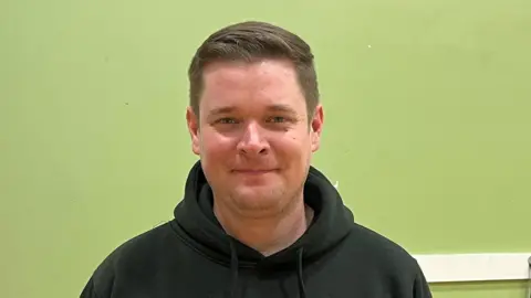 Benjamin Gillham has short brown hair and is wearing a black hooded top with a school logo and the word staff written on it. He is stood in a room with a green painted wall and microphone