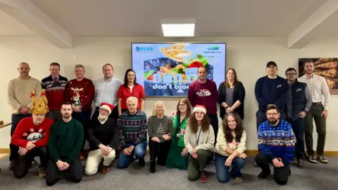 South West Water and ECAS staff pose for a photo. Some of the people are wearing Christmas jumpers and hats. A presentation slide is on a screen behind the people talking about what people should and should not put down their sink.