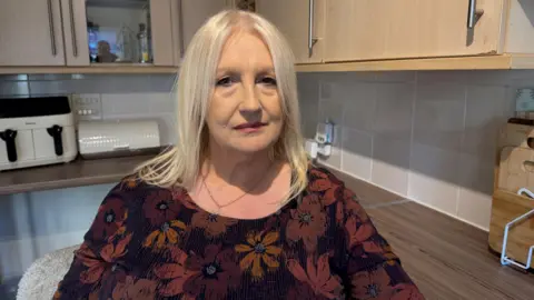 Julie Collett sitting in a kitchen. She is on older woman with straight grey hair. She has a smart red top on with flowers on and a necklace. Behind her we can see cupboards, a bread bin, an air fryer and chopping boards. 