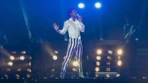 A film still showing a young man striking a pose on stage as he sings into a microphone. He has black hair in a mullet cut and wears a bright white shirt and white and blue striped trousers, which make him stand out against a dark background. The lighting is low and the shapes of his backing musicians can only just be made out. Stage lights shine in the background.