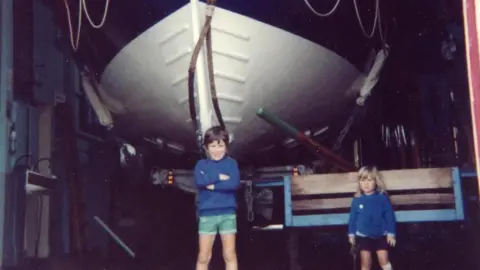 Karl Williams A boy with his arms crossed underneath a large boat, which is on a trailer and in a shed. A small girl is also in the picture, which appears to be quite old.