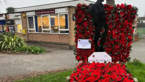 A poppy display placed outside a community centre, with a plaque stating 'Lest we forget'