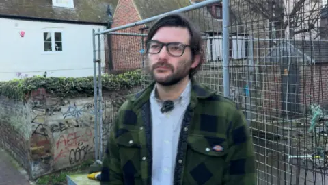 BBC/Phil Harrison A man with dark brown hair, glasses and a beard. He is wearing a green and black checked jacket and a light-blue shirt. He is standing in front of a fence. 