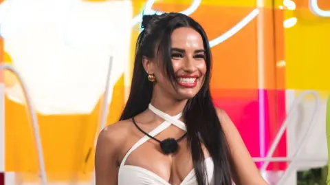 Ben Symons/Peacock via Getty Images Yulissa Escobar in the Love Island USA villa. Yulissa has long, straight, dark hair which she's styled in a half-up-half-down bun. She wears a strappy white dress and has a microphone around her neck. She smiles widely, wearing a red lipstick. The villa behind her is decorated with orange, yellow and pink.