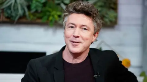 Aidan Gillen looks into the camera. He has brown scruffy hair with some white at the side. He is wearing a black jacket and dark round neck jumper.