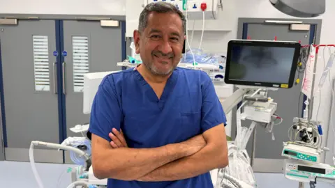 Handout Surgeon stands in operating theatre smiling, with his arms folded. He is wearing blue theatre scrubs. 