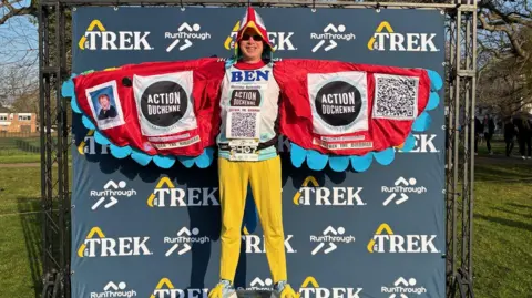 A man dressed in a parrot costume, standing on a small podium in front of a navy blue backdrop at a running race. The costume has red wings with blue feathers on their ends, and a hood that is red, with white around the eyes and a yellow beak. His trousers are yellow. Inside the wings, which are spread, there are two large logos for Action Duchenne. On the right is a black and white QR code and on the left is a photo of a young boy