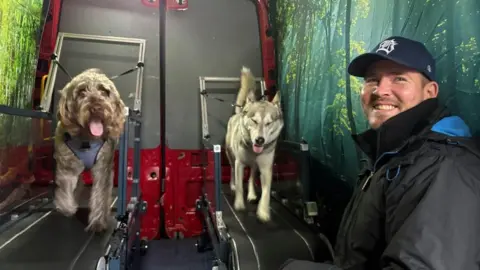 A man wearing a black rain coat and a baseball cap smiles at the camera. Behind him are two dogs running on two treadmills. The dog on the right is a grey husky type and the one on the left is a caramel coloured Poodle cross Labrador. They are all inside a red transit van, with large forest images on the inside walls.