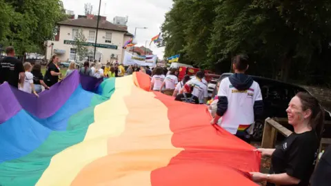 Doncaster Pride/Malcolm Johnston People holding a long rainbow flag stretched horizontally between them as they parade through a Doncaster street