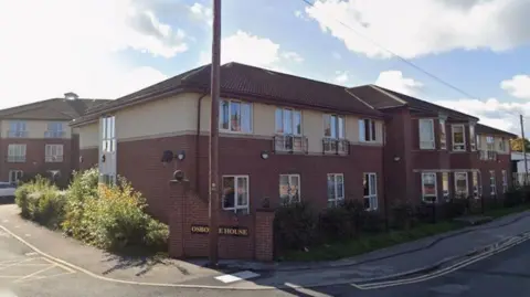 Google A two-storey brick building with cream-coloured upper walls and a brown tiled roof, identified as Osborne House by a sign near the entrance. The structure has multiple windows, some with small balconies, and is surrounded by low shrubs and greenery.