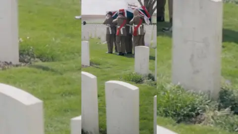 Men in British services uniforms carry a coffin, draped in the Union Jack, through a war cemetery.