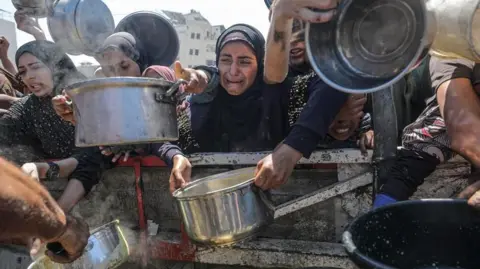 Mohammed Saber/EPA Várias pessoas segurando panelas para se encher de comida em um local de distribuição de ajuda - com uma mulher no meio vestindo todo preto visivelmente chateado e chorando