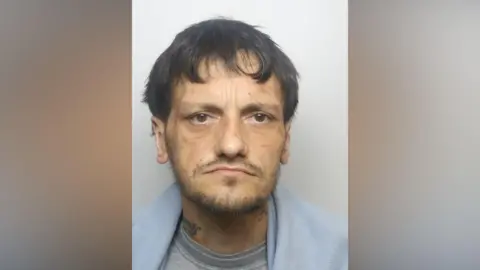 West Yorkshire Police A man with a gaunt face, brown hair and short stubble. He is pictured in a police mugshot with a blue jumper around his shoulders.