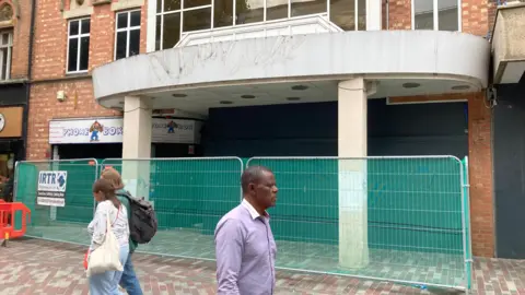 Alex Pope/BBC Three people walking past a shopping centre that is closed. There is a metal fence outside it, with netting on the fence.