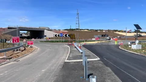 A newly constructed road junction and roundabout with new tarmac laid leading towards an underpass beneath a bridge, with road signs, traffic cones and temporary barriers. Workers in high-vis are standing near the junction, while earthworks, construction machinery and embankments are visible in the background under a clear blue sky.