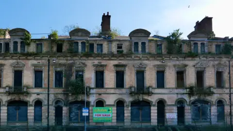 Close up image of the Marine Hotel. It is a large light brown brick building with greenery growing out of many of the windows.  On the fence surrounding the building is a green 'for sale' sign. 